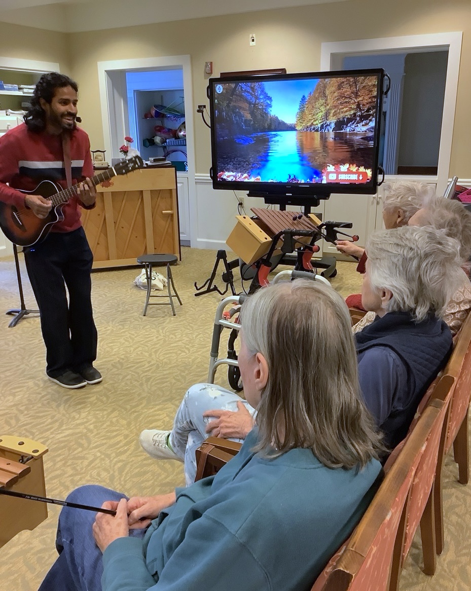 A music therapist playing guitar and singing for a group of older adults seated with mallet percussion instruments