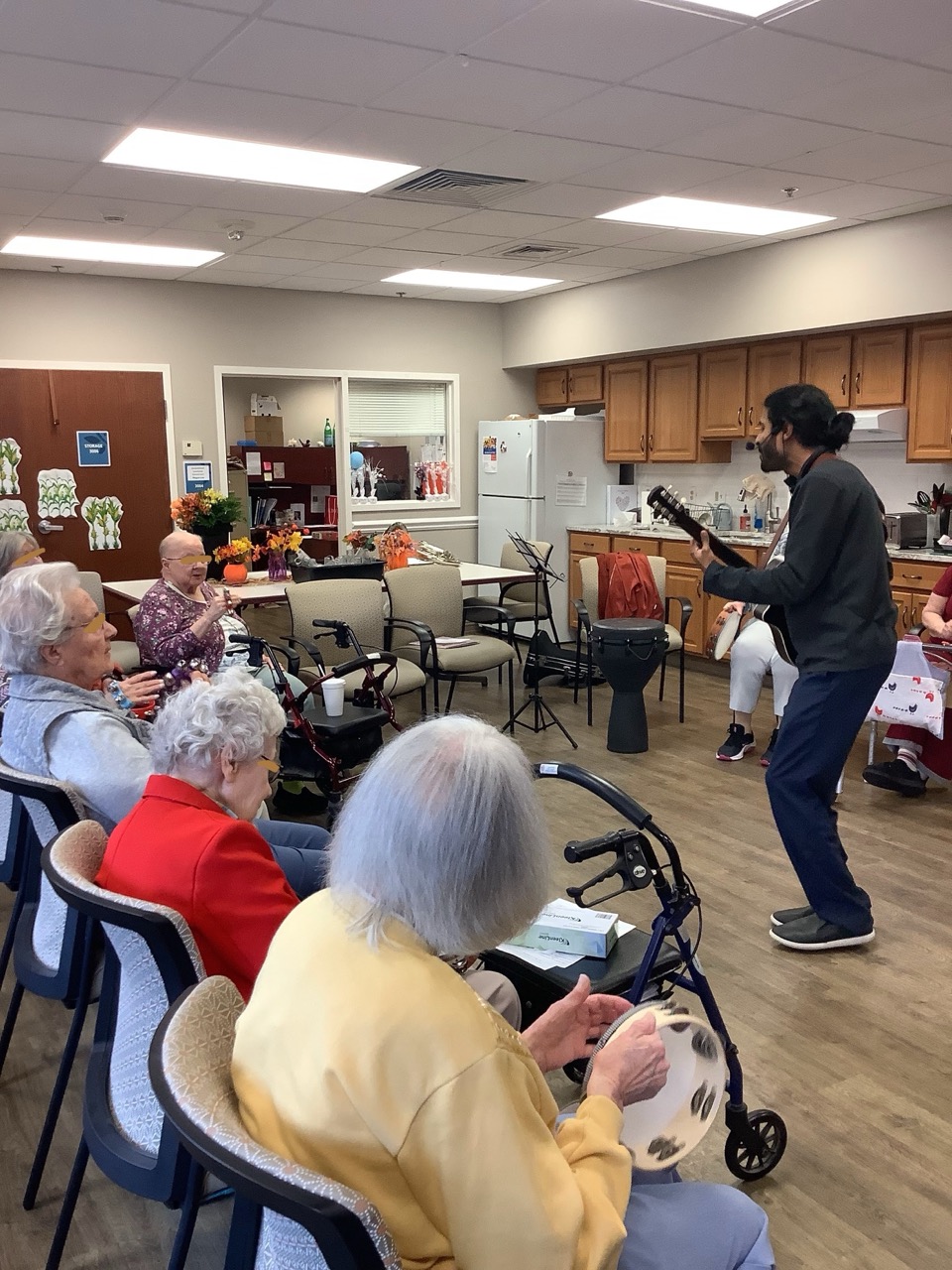 A music therapist playing guitar in a common room for older adults playing tambourines and a hand drum