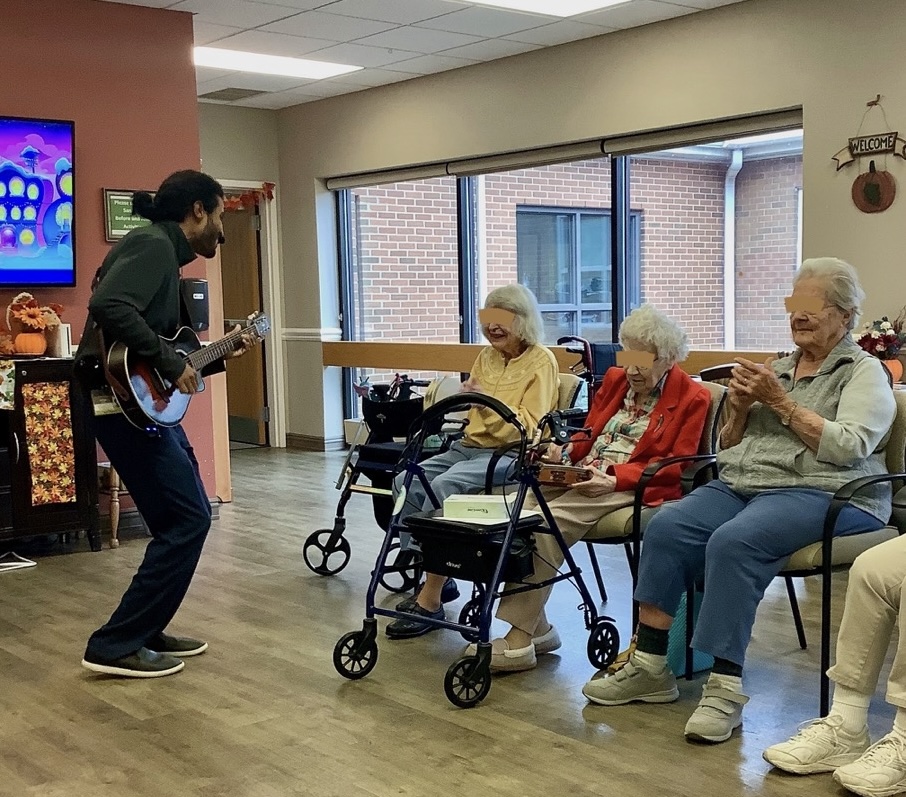 A music therapist playing guitar for a small group of older adults seated in a community room, smiling and clapping along