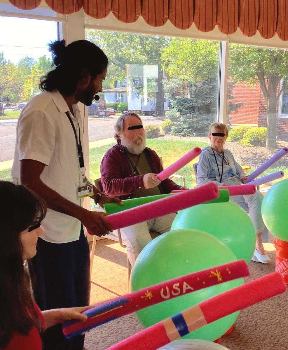 A music therapist leading a movement activity with pool noodles and exercise balls for a group of older adults